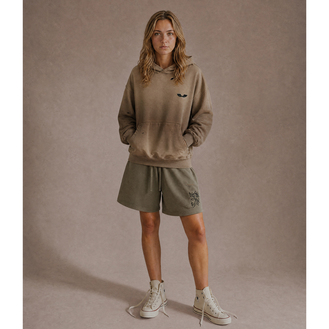 Full-body front lifestyle model view featuring the Foundations Series Heavyweight Unisex Sweat Short in artichoke green paired with the Crown of Thorns Heavyweight Hoodie in faded and distressed camel. Displays the high-density Christ Half Face leg embroidery, an oversized right-above-the-knee cut, and a boxy, hand-distressed dropped-shoulder hoodie silhouette for a complete premium streetwear look.