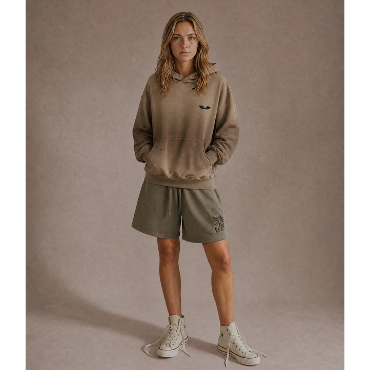 Full-body front lifestyle model view featuring the Foundations Series Heavyweight Unisex Sweat Short in artichoke green paired with the Crown of Thorns Heavyweight Hoodie in faded and distressed camel. Displays the high-density Christ Half Face leg embroidery, an oversized right-above-the-knee cut, and a boxy, hand-distressed dropped-shoulder hoodie silhouette for a complete premium streetwear look.