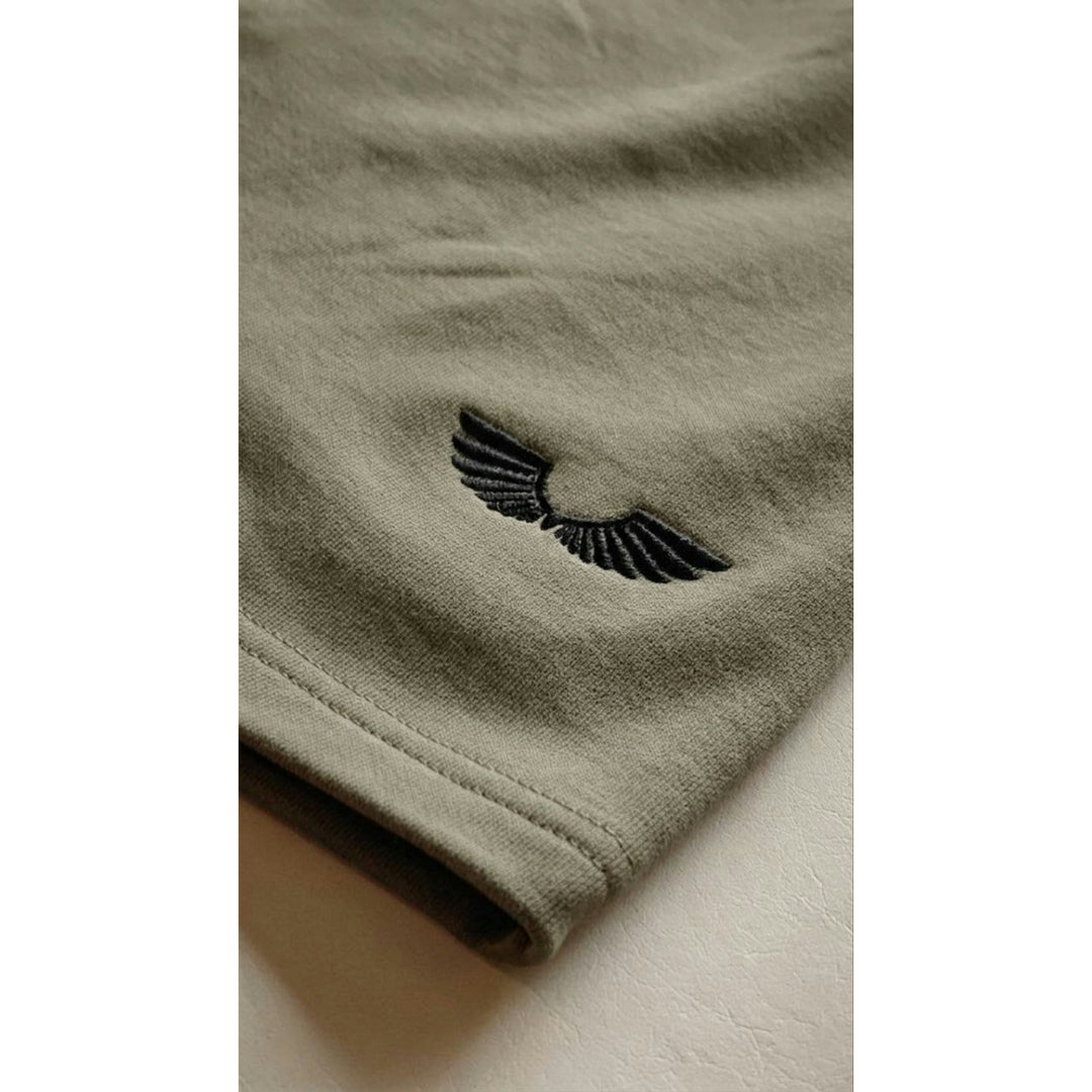 Macro detailing shot of the Foundations Series Heavyweight Unisex Sweat Short in artichoke green. Close-up highlights the signature Apostles Apparel black embroidered wings logo on the rear right leg cuff, showcasing the premium high-density stitching, durable coverstitch cuff hem, and tight weave of the 425GSM combed cotton French terry fabric.
