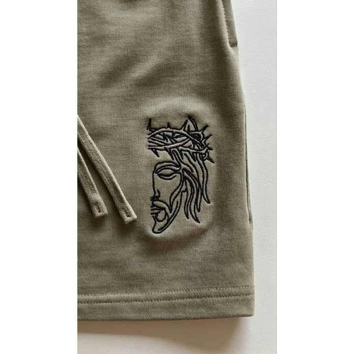 Macro detailing shot of the Foundations Series Christian Heavyweight Unisex Sweat Short in artichoke green. Close-up highlights the premium high-density "Christ Half Face" line-art embroidery on the front leg panel, showcasing the intricate black stitch pattern against the dense texture of the 425GSM combed cotton French terry fabric.