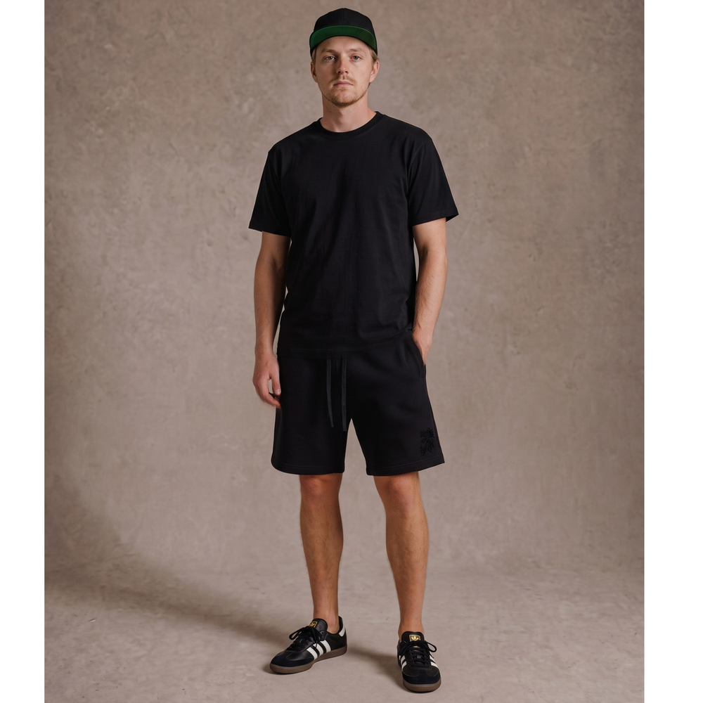 Full-body lifestyle model view wearing the Apostles Apparel Foundations Heavyweight Unisex Sweat Short in black. Showcases the relaxed oversized streetwear fit cutting right above the knee, paired with a matching black tee, featuring the subtle tonal black high-density Christ Half Face embroidery on the left front leg and an overbuilt drawcord waistband.