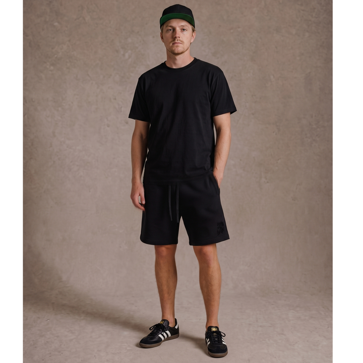 Full-body lifestyle model view wearing the Apostles Apparel Foundations Heavyweight Unisex Sweat Short in black. Showcases the relaxed oversized streetwear fit cutting right above the knee, paired with a matching black tee, featuring the subtle tonal black high-density Christ Half Face embroidery on the left front leg and an overbuilt drawcord waistband.