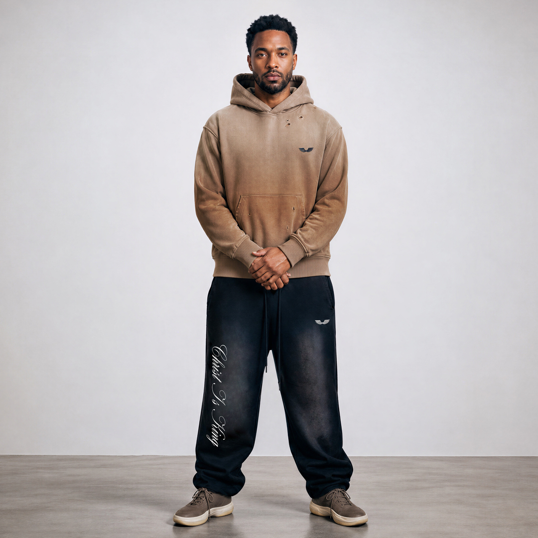Full-body front lifestyle model view featuring the Apostles Apparel Foundations Christ Is King Sun-Faded Heavyweight Pant in black paired with the Crown of Thorns Heavyweight Hoodie in faded camel. Showcases the relaxed oversized tapered jogger fit with right-leg vertical calligraphy, combined with the boxy dropped-shoulder hoodie silhouette for a complete premium Christian streetwear outfit look.