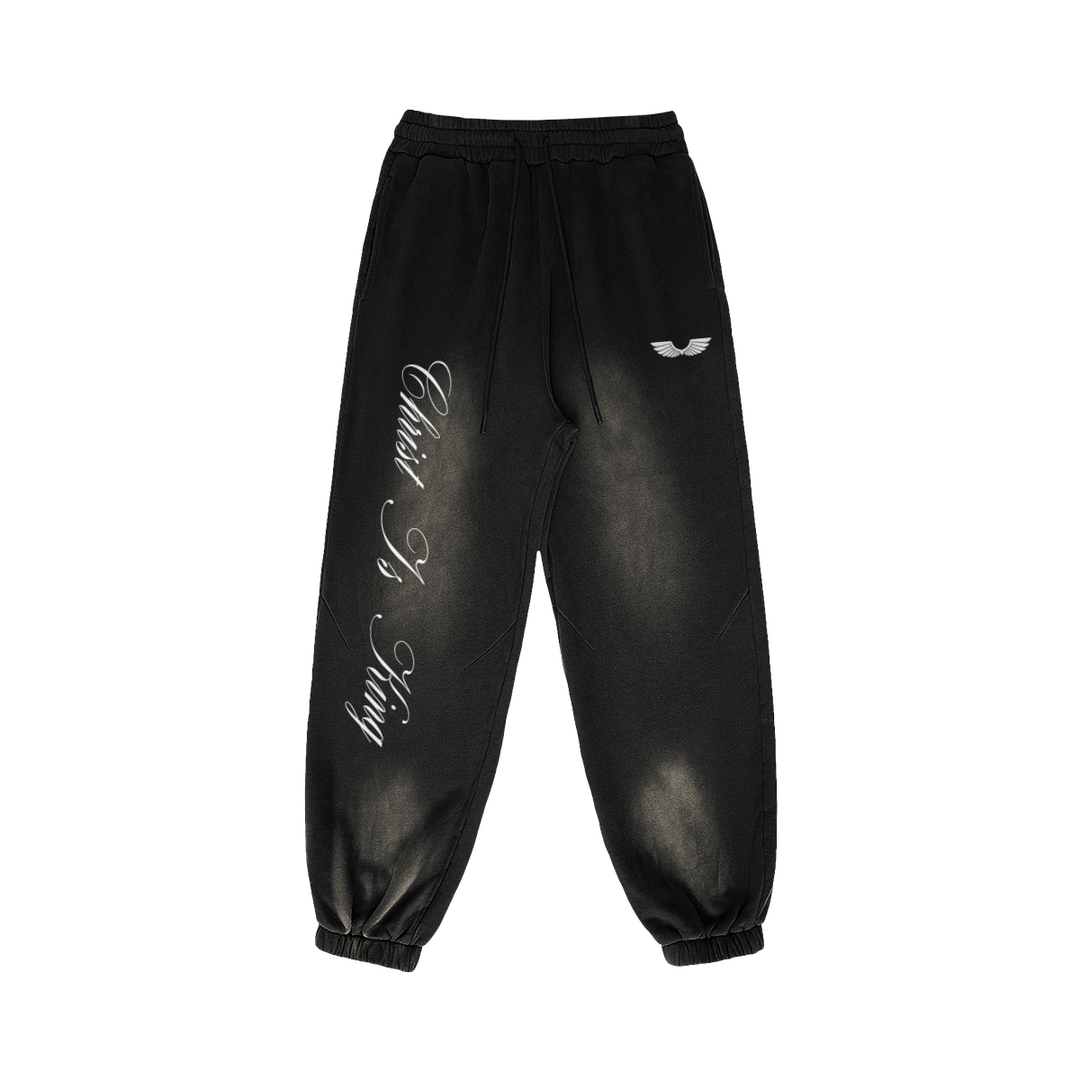 Front studio flat lay view of the Foundations Series Christ Is King Sun-Faded Heavyweight Pant in a smokey black tone. Features crisp white calligraphy down the right leg, a premium embroidered brand wings logo on the left hip, a structural 3-tiered topstitched waistband with thick drawcords, and gathered ankle cuffs showcasing the 400GSM combed cotton relaxed tapered silhouette.