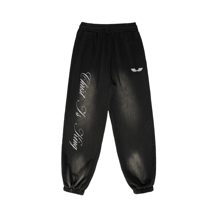 Front studio flat lay view of the Foundations Series Christ Is King Sun-Faded Heavyweight Pant in a smokey black tone. Features crisp white calligraphy down the right leg, a premium embroidered brand wings logo on the left hip, a structural 3-tiered topstitched waistband with thick drawcords, and gathered ankle cuffs showcasing the 400GSM combed cotton relaxed tapered silhouette.