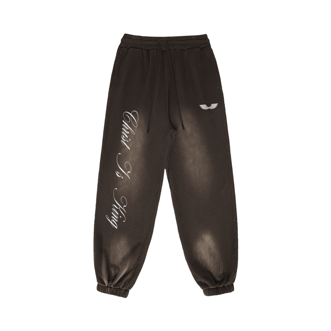 Front studio flat lay view of the Foundations Series Christ Is King Sun-Faded Heavyweight Pant in a smokey brown tone. Features sharp white calligraphy down the right leg, a premium embroidered brand wings logo on the left hip, a structural 3-tiered topstitched waistband with thick drawcords, and gathered ankle cuffs showcasing the 400GSM combed cotton relaxed tapered silhouette.