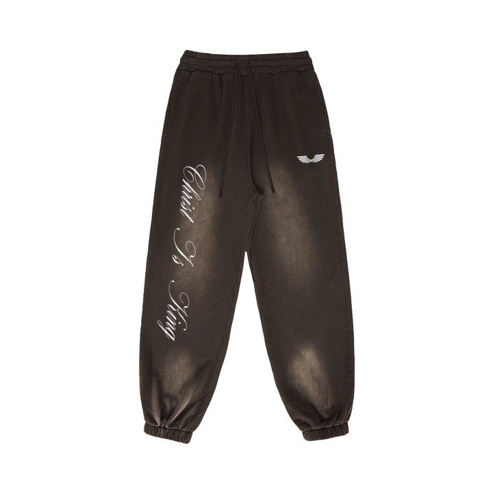 Front studio flat lay view of the Foundations Series Christ Is King Sun-Faded Heavyweight Pant in a smokey brown tone. Features sharp white calligraphy down the right leg, a premium embroidered brand wings logo on the left hip, a structural 3-tiered topstitched waistband with thick drawcords, and gathered ankle cuffs showcasing the 400GSM combed cotton relaxed tapered silhouette.