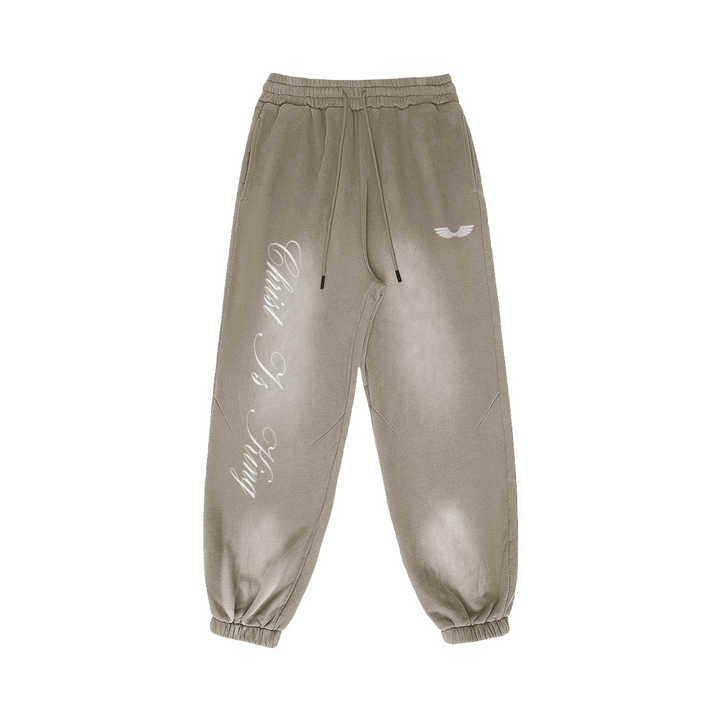 Front studio flat lay view of the Foundations Series Christ Is King Sun-Faded Heavyweight Pant in a smoky grey tone. Features razor-sharp vertical calligraphy down the right leg, a premium stitched brand wings logo on the left hip, an architectural 3-tiered topstitched waistband with heavy drawcords, and elasticized ankle cuffs showcasing the 400GSM combed cotton tapered silhouette.