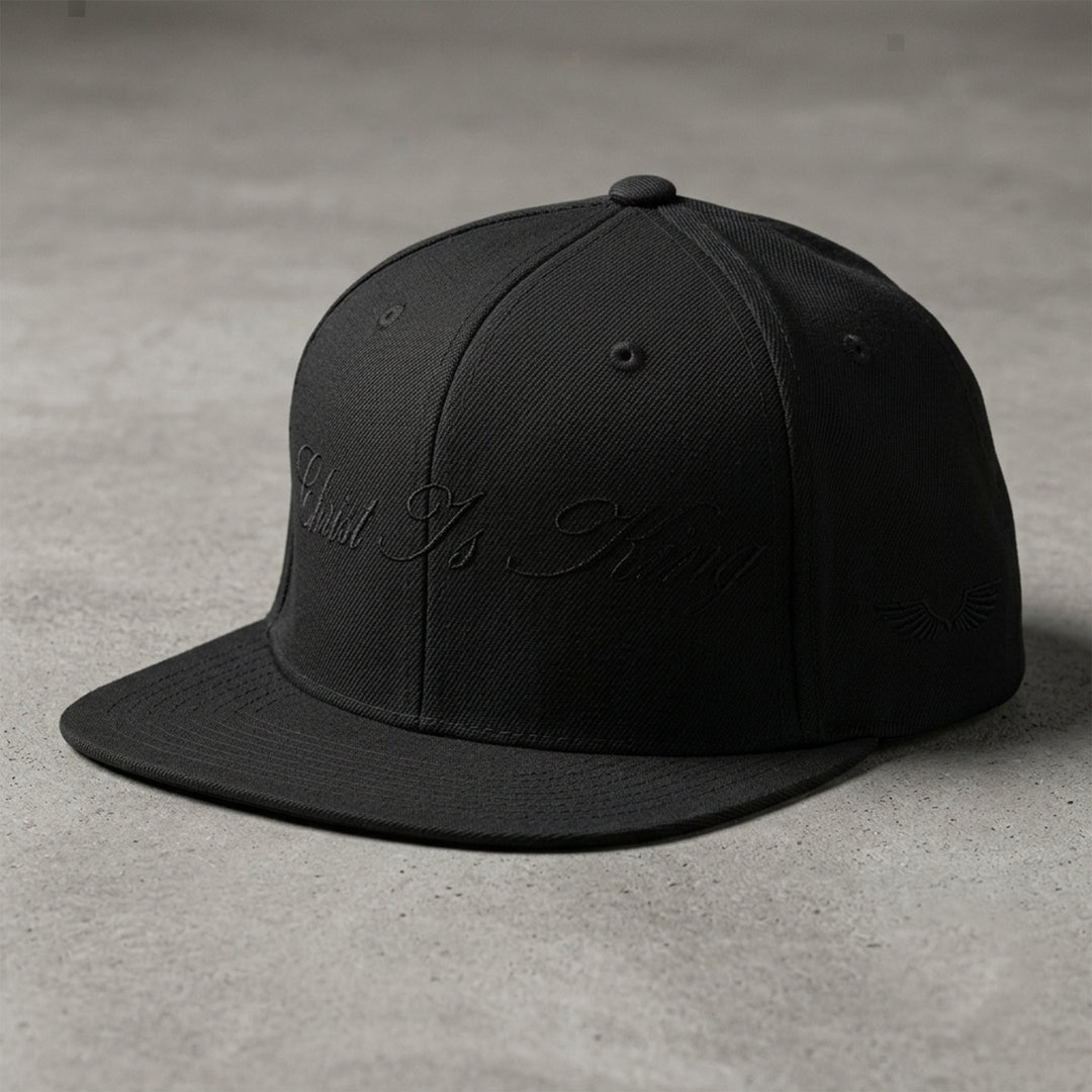 Studio view of the Foundations Series Christ Is King Triple Black Signature Snapback by Apostles Apparel. Features a structured 6-panel high-profile crown silhouette, a tonal black flat-stitch calligraphy design on the front panel, and side wings embroidery on a premium heavyweight acrylic and wool blend chassis.