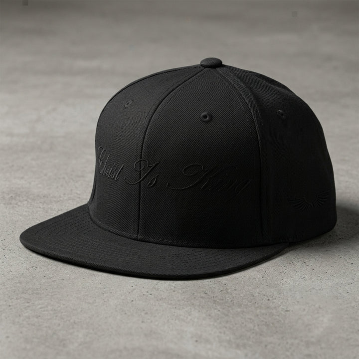 Studio view of the Foundations Series Christ Is King Triple Black Signature Snapback by Apostles Apparel. Features a structured 6-panel high-profile crown silhouette, a tonal black flat-stitch calligraphy design on the front panel, and side wings embroidery on a premium heavyweight acrylic and wool blend chassis.