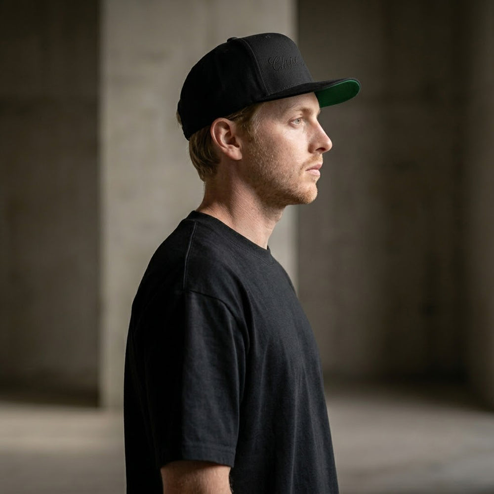 Side profile model shot wearing the Apostles Apparel Christ Is King Triple Black Signature Snapback. Highlights the classic throwback green undervisor accent, high-profile structured 6-panel crown build, flat visor bill, and tonal black calligraphy flat-stitch embroidery details.