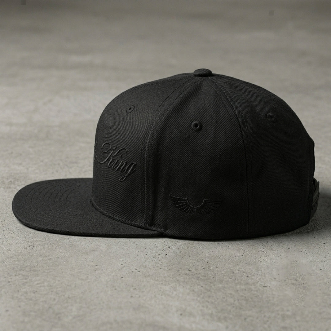 Side studio view of the Foundations Series Christ Is King Triple Black Signature Snapback by Apostles Apparel on a concrete surface. Showcases the tonal black embroidered wings brand logo on the side panel, the rigid high-profile 6-panel structure, and premium heavyweight acrylic-wool fabric.