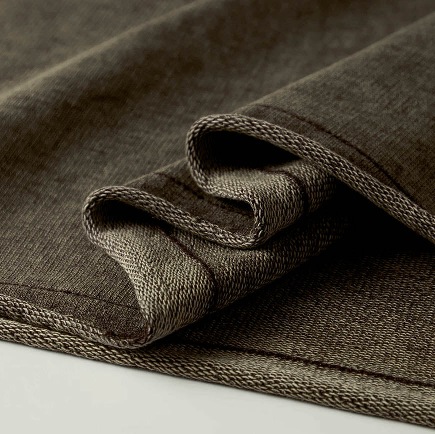 Macro detailing shot of the Foundations Series 70X7 Raw Hem Long Sleeve in a sun-faded black tone. Close-up tightly isolates the character-driven aged raw-edge hem finish, highlighting the structural multi-dimensional washed texture and dense grain of the heavy 260GSM enzyme-washed combed cotton fabric for a premium Christian apparel statement piece.