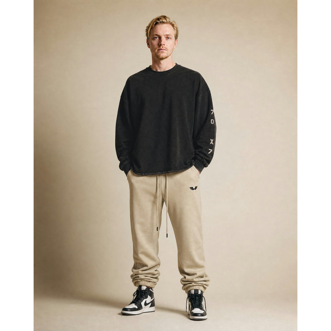 Full-body studio lifestyle model view featuring the Foundations Series 70X7 Raw Hem Long Sleeve in a sun-faded black tone paired with the Heavyweight Unisex Brushed Fleece Jogger in a camel tone. Showcases the boxy, drop-shoulder long-sleeve silhouette with its weathered vertical stencil 70X7 sleeve design alongside the relaxed tapered jogger stacking naturally over high-top sneakers, delivering a premium Christian streetwear outfit choice for mission-driven Christian clothing and pro-life apparel.