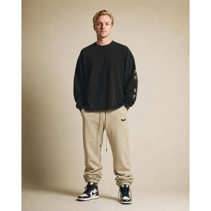 Full-body studio lifestyle model view featuring the Foundations Series 70X7 Raw Hem Long Sleeve in a sun-faded black tone paired with the Heavyweight Unisex Brushed Fleece Jogger in a camel tone. Showcases the boxy, drop-shoulder long-sleeve silhouette with its weathered vertical stencil 70X7 sleeve design alongside the relaxed tapered jogger stacking naturally over high-top sneakers, delivering a premium Christian streetwear outfit choice for mission-driven Christian clothing and pro-life apparel.