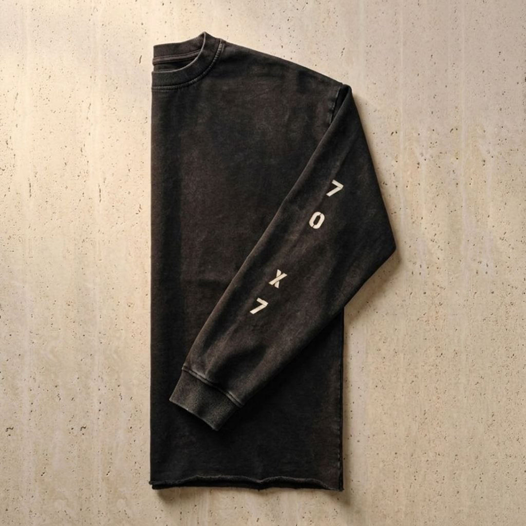 Side-folded studio flat lay view of the Foundations Series 70X7 Raw Hem Long Sleeve in a sun-faded black tone. Tightly showcases the weathered vertical stencil 70X7 graphic down the sleeve panel, the robust double-stitched ribbed collar, and the distinctive character-driven aged raw-edge hem finish on a heavy 260GSM combed cotton canvas for premium Christian streetwear.