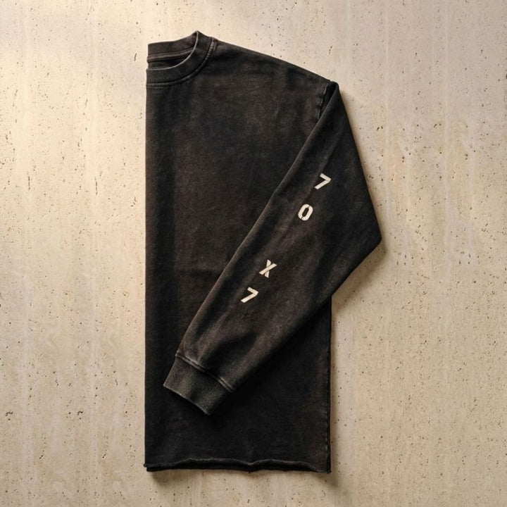 Side-folded studio flat lay view of the Foundations Series 70X7 Raw Hem Long Sleeve in a sun-faded black tone. Tightly showcases the weathered vertical stencil 70X7 graphic down the sleeve panel, the robust double-stitched ribbed collar, and the distinctive character-driven aged raw-edge hem finish on a heavy 260GSM combed cotton canvas for premium Christian streetwear.