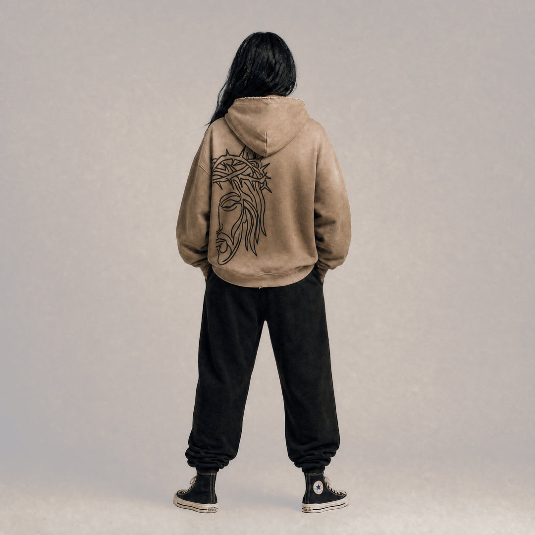 Full-body lifestyle model view from the back wearing the Apostles Apparel Foundations Crown of Thorns Heavyweight Hoodie in faded and distressed camel. Displays the signature boxy oversized silhouette with dropped shoulders, highlighting the raw streetwear drape and the jumbo line-art back screen print.