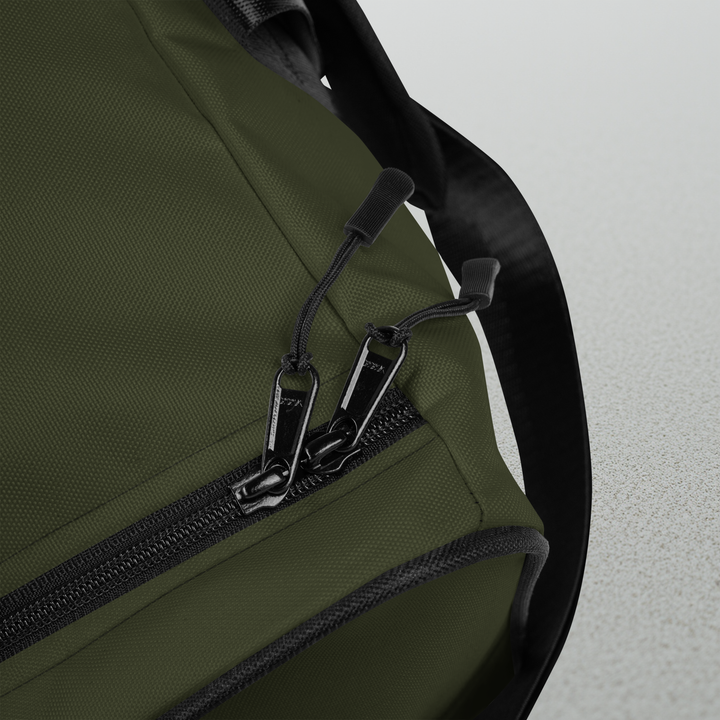 Macro detailing shot of the Foundations Series Let God Carry It Utility Duffle Bag in olive green. Close-up highlights the heavy-duty dual zipper pulls on the main compartment, the rugged texture of the 9 oz./yd.² high-density polyester chassis, and the clean finish of the reinforced black T-piping architecture.