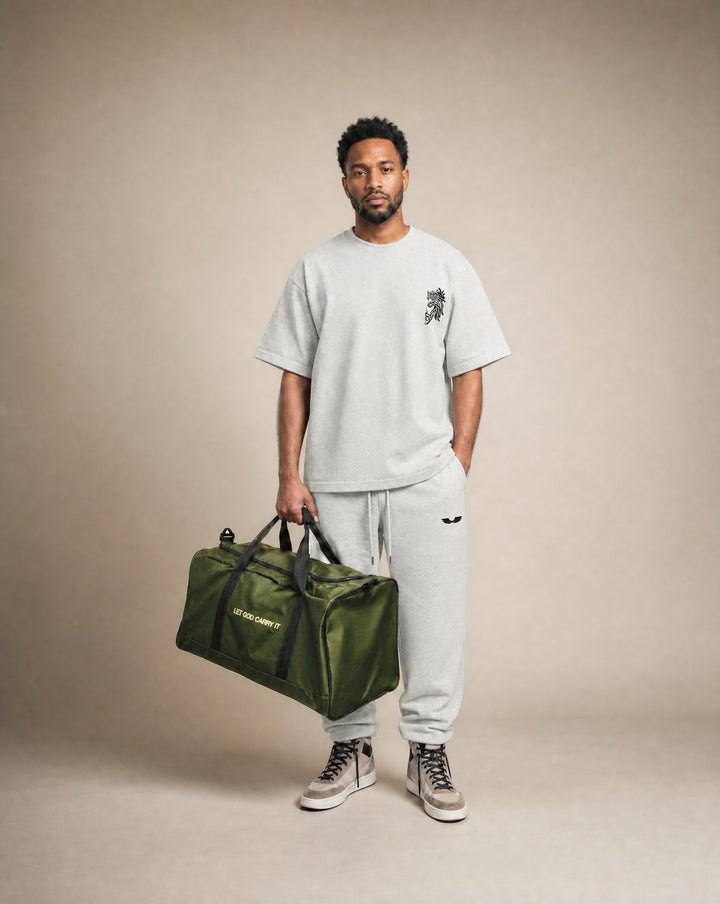 Studio model shot featuring the Apostles Apparel Foundations Let God Carry It Utility Duffle Bag in olive green. The model holds the bag by its dual padded mesh handles, showcasing the true 50-liter capacity scale and rigid reinforced T-piping architecture.