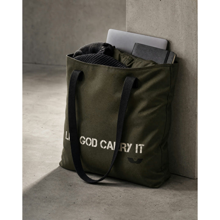 Product context shot of the Foundations Series Let God Carry It Utility Tote Bag in olive green leaning against a concrete pillar. Displays the bag loaded with a laptop and notebook, showcasing the structural stability, 10L capacity, and durable 44-pound weight limit chassis.