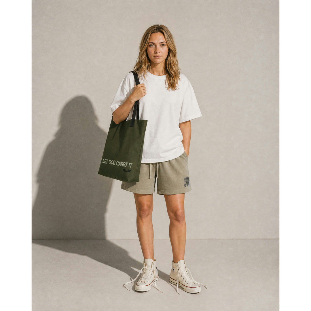 Full-body lifestyle model shot featuring the Apostles Apparel Foundations Let God Carry It Utility Tote Bag in olive green. Showcases the 26-inch natural cotton bull denim handles worn over the shoulder, highlighting the scale of the 15x15 high-capacity streetwear silhouette.