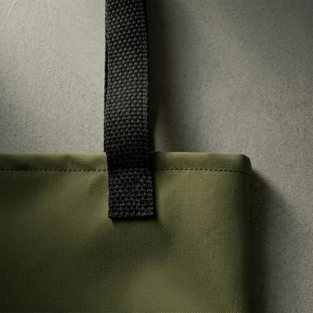 Macro detailing shot of the Foundations Series Let God Carry It Utility Tote Bag in olive green. Close-up highlights the industrial-grade rugged construction, showcasing the dense weave of the high-density polyester chassis and the secure, reinforced stitching of the black cotton bull denim dual handles.